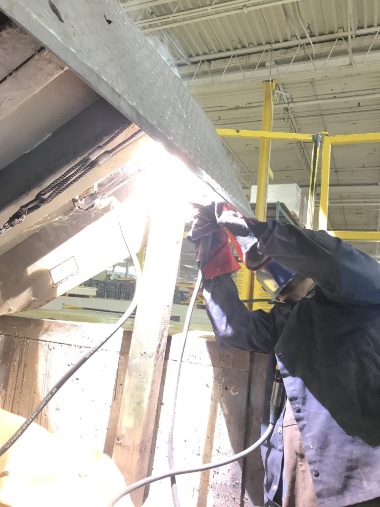 The welder welds hinges on the lip of misaligned loading dock. The dislocation is eliminated by a came along with the sling correcting the misalignment. JW Portable Welding, London, Ontario, 2021