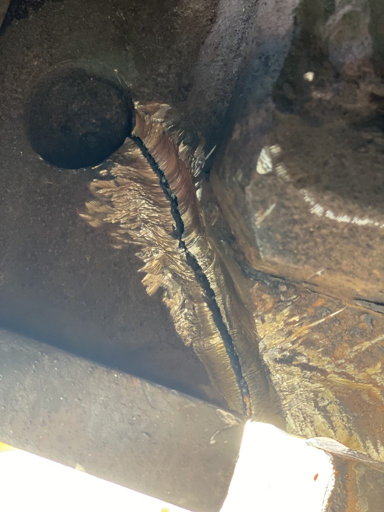 The crack in 1 inch steel connects the edge of the bumper of locomotive for the railway crane with the bore of about 2 inch, The grooving and consequently welding had taken place in extremely difficult position laying down on railway tracks. The groove depth is about 0.5 inch. The crack development represents an engineering failure due to accumulation of stress between the rim of 2 inch thick and the bore. London ON, 2024 for JW Portable Welding & Repairs.