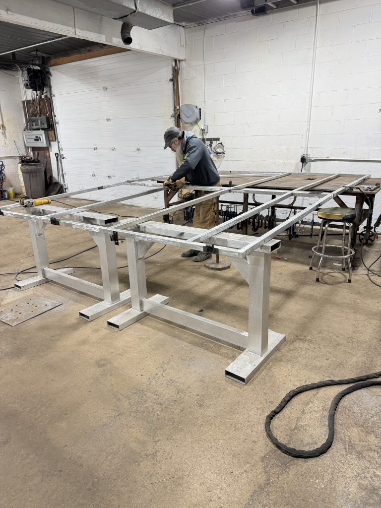 Aluminum fabrication of a huge sign frame structure. The sign take the whole table 4 feet x 12 feet , support in the middle and two aluminum support horses. Total measurements of sign in this position is 16 feet x 10 feet. Such fabrication requires large space with multiple supports. For JW Portable Welding, 2026, Shop in Lucan, Ontario